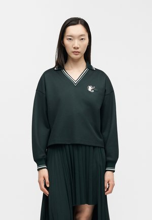 Jonge vrouw die een donkergroen poloshirt met lange mouwen en witte strepen en een bijpassende geplooide rok draagt, kijkt recht vooruit tegen een effen achtergrond.