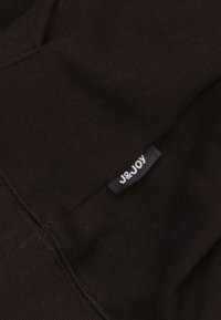 Zwarte stof met een gladde textuur. Bevat een J&Joy-label dat aan de rand is gestikt. Eenvoudig ontwerp met zichtbare stikdetails.