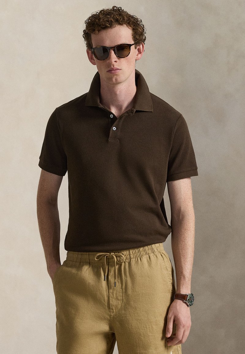 Brown polo shirt with a textured finish, three-button placket, and short sleeves, paired with beige drawstring shorts and sunglasses.