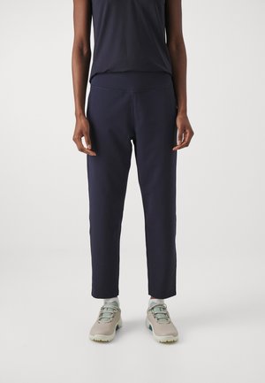 Personne portant un pantalon droit bleu marine foncé, des baskets claires avec des lacets vert clair, et un haut sans manches foncé sur un fond uni.