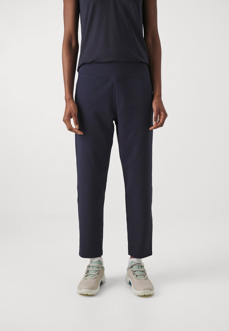 Personne portant un pantalon droit bleu marine foncé, des baskets claires avec des lacets vert clair, et un haut sans manches foncé sur un fond uni.