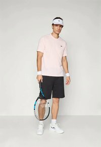 Noor mees, kes kannab helesinist särki, musti lühikesi pükse, valget visiitäit, randme- ja tossusid, hoiab tennisereketit, seistes ühtlase tausta ees.