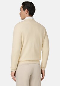 Kremowy sweter z dzianiny z prążkowanymi mankietami i dolnym brzegiem, pokazany od tyłu, w połączeniu z jasnobeżowymi spodniami. Miękka faktura i proste detale wzornicze.