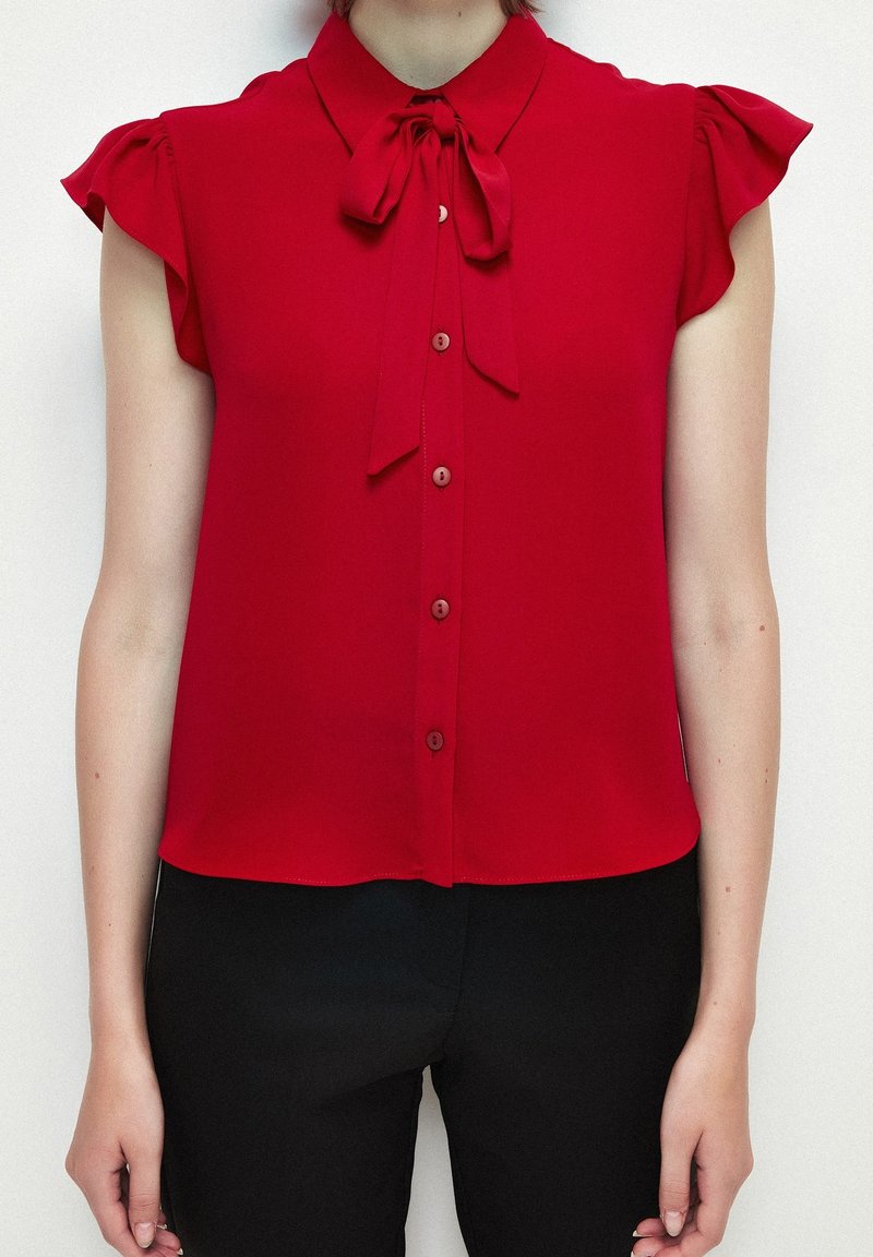 Femme portant un haut sans manches rouge avec un col en nœud papillon et des boutons sur le devant, associé à un pantalon noir taille haute.