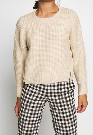 Femme portant un pull en maille texturée crème avec des fermetures éclair latérales et un pantalon à carreaux noir et blanc, debout devant un fond uni.