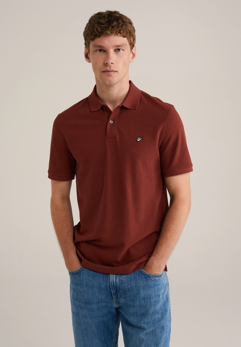 Mørkerød polo skjorte, korte ærmer, med en tre-knaps placket. Har et lille broderet logo på venstre bryst. Båret med blå jeans.