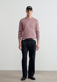 Νέος άνδρας που στέκεται φορώντας μια ρόδινη πουλόβερ με σκόνη, μαύρο παντελόνι, μαύρη ζώνη, μαύρα παπούτσια και μαύρο καπέλο μπροστά σε έναν απλό λευκό τοίχο.