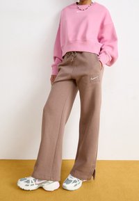 Rosa kortärmad sweatshirt med ribbade manschetter, ihop med bruna sweatpants med sidofickor och en liten logotyp. Vita och grå sneakers.