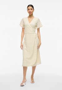Kietaisutunika vaaleasta beige pitsistä, jossa on lyhyet, löysät hihat ja vyötäröllä solmittava nauha. Sisältää tekstuuripinnan ja ulottuu puoliväliin pohkeita.