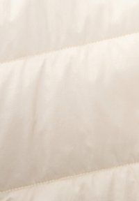 Tissu matelassé beige avec une texture lisse, présentant une couture diagonale. La surface semble douce et légèrement brillante.