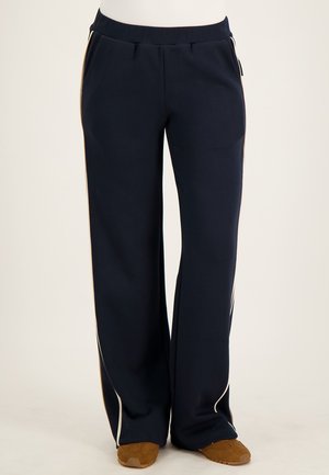 Marineblauwe wijde broek met witte zijstrepen, elastische taille, voorzakken, gedragen met bruine schoenen op een neutrale achtergrond.