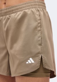 Shorts de sport beige avec ceinture élastique, poche latérale et logo Adidas blanc sur la cuisse avant gauche au-dessus du short de compression intégré.