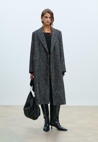 Cappotto lungo a fantasia houndstooth bianco e nero, con design doppiopetto, ampi rever e tasche laterali. Indossato con stivali neri al ginocchio.