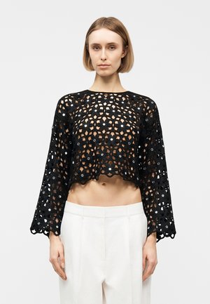 Kobieta ubrana w czarny, kwiecisty, koronkowy top z długim rękawem i białe spodnie z wysokim stanem, stojąca na tle jasnego, jednolitego tła.