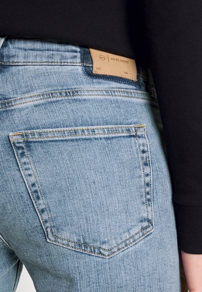 Jeans en denim bleu clair avec poche arrière et étiquette en cuir beige portant la mention "AG-ED DENIM", portés avec un haut noir.