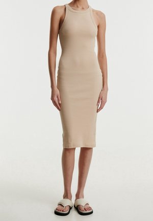 Mujer con vestido midi sin mangas beige con textura de canalé y sandalias deslizantes a juego, de pie frente a un fondo claro y liso.
