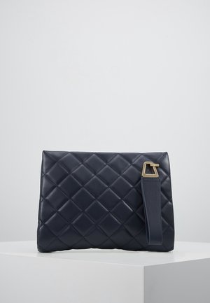 Laivastonsininen tikattu nahkainen clutch-laukku, jossa leveä hihna ja kullanvärinen kolmionmuotoinen metallilukko, valkoisen jalustan päällä.