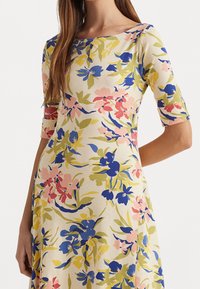 Blommig klänning i krämfärgad tyg; har blå, rosa och gröna blommor; båtutskuren halsringning och ärmar i armbågslängd; flödande A-linjeform.