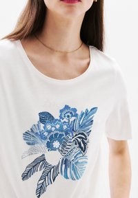 Witte t-shirt met een blauw bloemontwerp met verschillende bladeren en bloemen. Korte mouwen, ronde halslijn, gladde textuur, eenvoudige pasvorm.