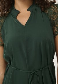 Tmavě zelené šaty s výstřihem do V a volánkovou lemovkou. Mají krátké krajkové rukávy a stažený pas s detailem na zavazování. Hladká textura.