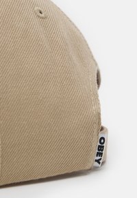 Boné de algodão bege com uma superfície texturizada. Apresenta uma etiqueta branca com "OBEY" em preto, uma costura bordada e um buraco de ventilação circular.