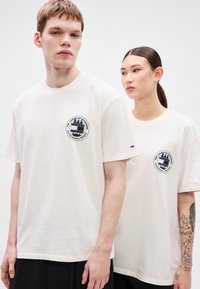 Bombažne majice v barvi slonokoščene kosti, kratkimi rokavi in okroglim izrezom. Na sprednji strani je na levi strani prsi okrogel črn grafični logotip.