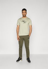 Helles olivengrünes Sport-T-Shirt mit einem schwarzen Logo und horizontalen Streifen, kombiniert mit dunkeloliven Jogginghosen und schwarzen Sneakers.