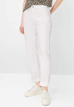 Donna che indossa pantaloni bianchi risvoltati, sneakers bianche e una maglia marrone a pois, in piedi davanti a uno sfondo chiaro e uniforme.