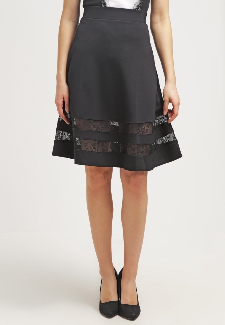 Femme portant une jupe noire longueur genou avec des panneaux de dentelle horizontaux près de l'ourlet et des talons hauts noirs fermés.