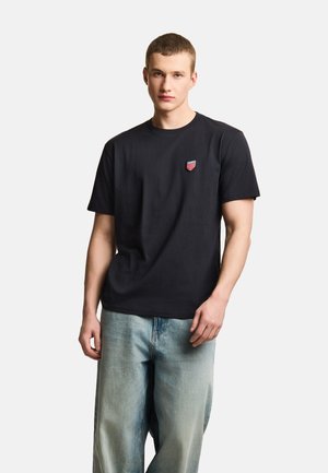 Młody mężczyzna stoi, ubrany w prostą czarną koszulkę z małym czerwono-białym emblematem oraz jasnoniebieskie, luźne dżinsy.