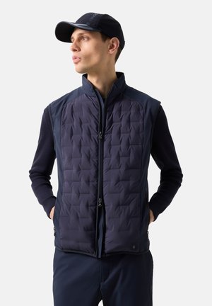 Fiatal férfi tengerészkék steppelt mellényben, hosszú ujjú inggel és hozzá illő sapkával, zsebbe tett kézzel áll, balra néz, egyszínű háttér előtt.