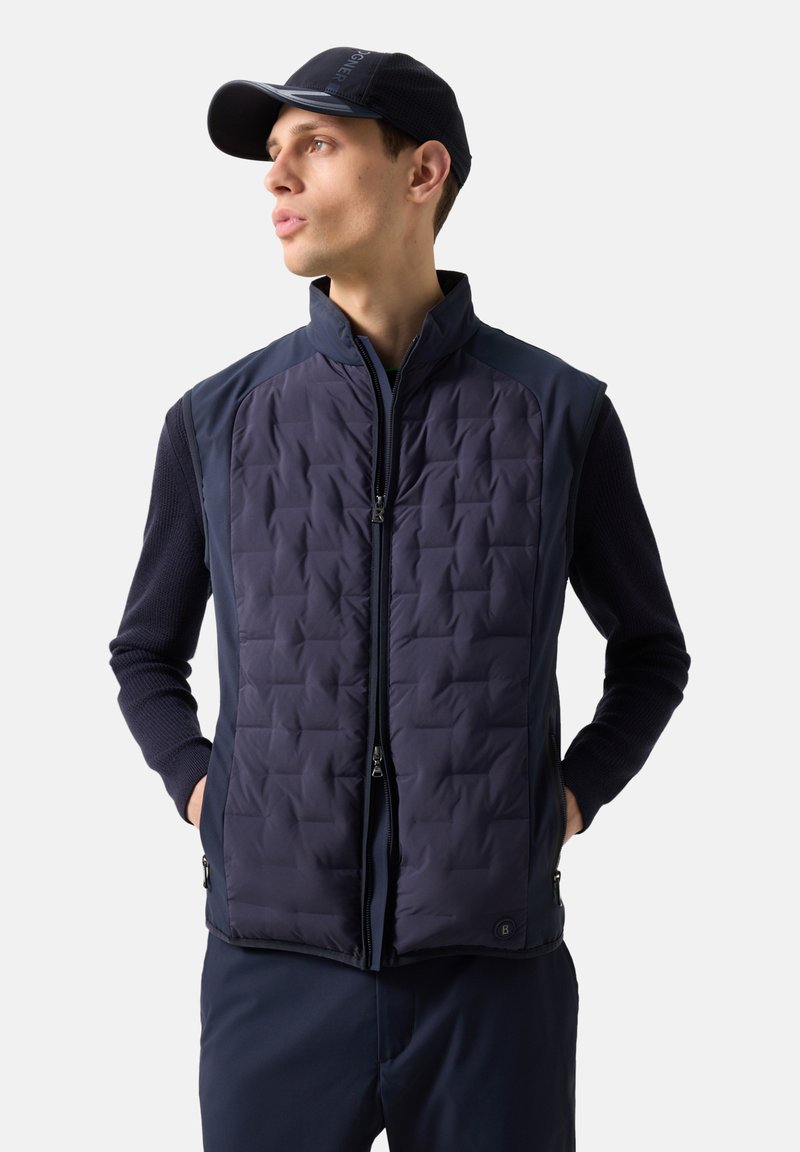 Νεαρός άνδρας που φοράει σκούρο μπλε καπιτονέ γιλέκο πάνω από μακρυμάνικο πουκάμισο και καπέλο που ταιριάζει, στέκεται με τα χέρια στις τσέπες, κοιτάζοντας αριστερά σε απλό φόντο.