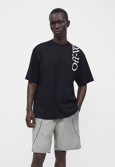 Άνδρας που φοράει μαύρο oversized T-shirt με λευκό κάθετο κείμενο στον ώμο και ανοιχτό γκρι σορτς με μαύρο περίγραμμα, στέκεται μπροστά σε απλό φόντο.