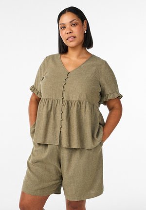Kvinde med glat sort hår iført olivengrøn knaplukket kortærmet top med flæser og matchende løstsiddende shorts, hænderne i lommerne.