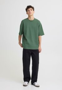 T-shirt vert surdimensionné à manches courtes en coton doux, associé à un pantalon noir coupe décontractée et des baskets blanches avec des accents gris.