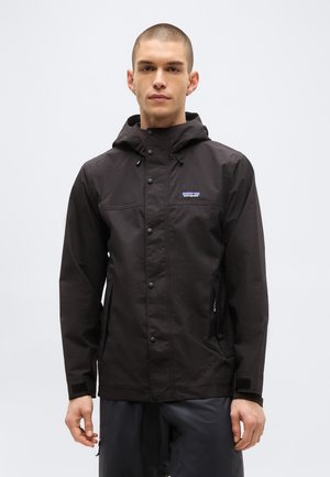 Fiatal férfi fekete kapucnis esőkabátot visel, patentokkal és oldalzsebekkel, tiszta világos háttér előtt áll.