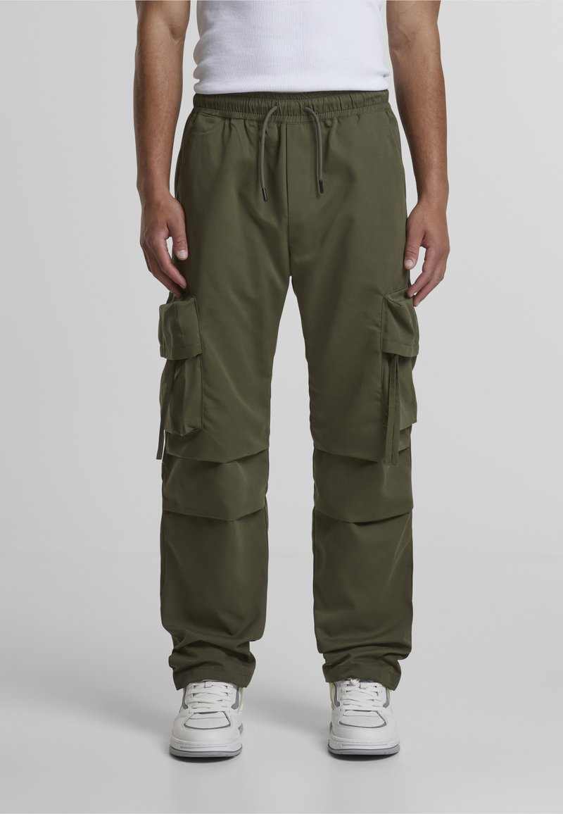 Persona que lleva pantalones cargo verde oliva con múltiples bolsillos y zapatillas blancas, de pie frente a un fondo liso.