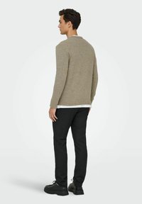 Světle hnědý pletený svetr s kulatým výstřihem, nošený přes bílou košili, v kombinaci s černými slim-fit kalhotami a černými teniskami.