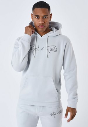 Sudadera gris clara hecha de tela suave, con un bolsillo tipo canguro y un logotipo bordado de "Project x Paris" en el pecho y la manga.
