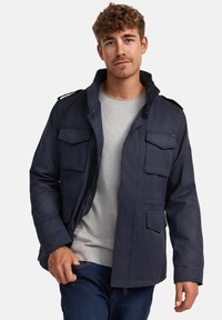 Marineblauwe utilityjack met meerdere voorzakken, ritssluiting en een hoge kraag. Draag het over een lichtgrijs shirt met een ontspannen pasvorm.