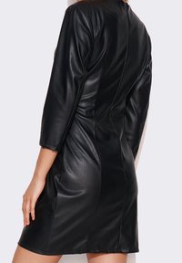 Donna che indossa un vestito attillato in ecopelle nera con maniche a tre quarti, vista da dietro su uno sfondo chiaro e semplice.