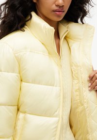 Femme aux cheveux ondulés portant une doudoune jaune pâle par-dessus un pull texturé jaune clair, tenant la veste ouverte d'une main.