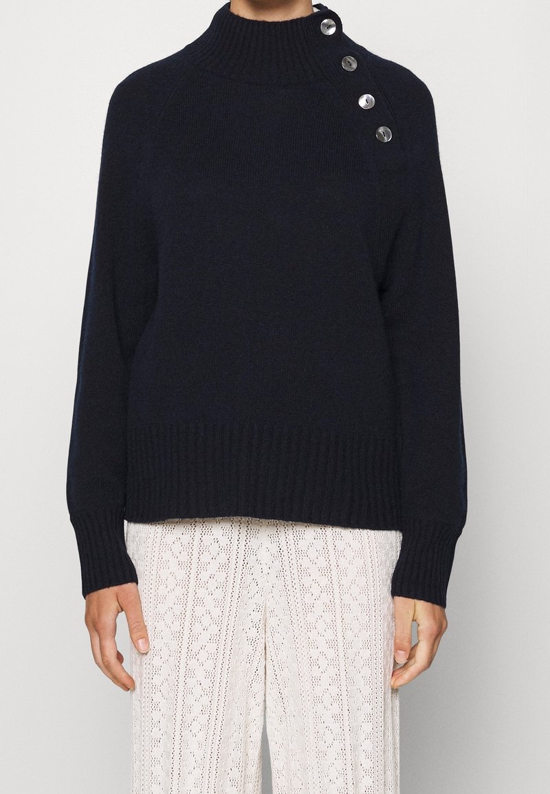 Pull en maille côtelée marine avec col montant et quatre boutons métalliques sur l'épaule, associé à un pantalon à jambes larges blanc à motifs.