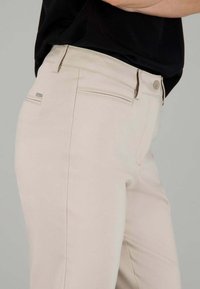 Pantaloni in misto cotone beige con tasche laterali e posteriori. Presentano una texture liscia, una vestibilità dritta e un piccolo logo metallico sul retro.