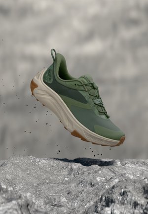 Chaussure de sport verte avec semelle blanche et marron flottant au-dessus d'un sol rocailleux avec de petites particules de terre en suspension dans l'air.