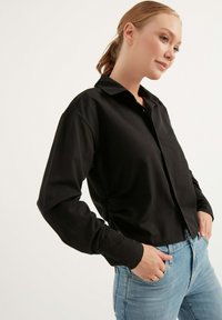 Schwarzes Langarmshirt mit Kragen, das eine figurbetonte Silhouette und geraffte Seitendetails aufweist. Glattes Material, kombiniert mit hellblauen Jeans.