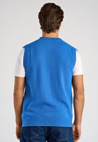 Blå stickad ärmlös tröja med ribbad halsringning och linning, med en mjuk textur. Bärs över en vit skjorta och kombineras med mörka jeans.