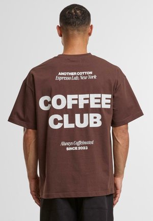 Brown cotton t-shirt featuring large white text: "COFFEE CLUB," with smaller text above and below, labeled "Another Cotton" and "Always Caffeinated Since 2023."