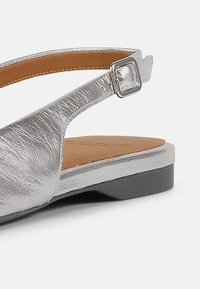 Sandalias metálicas plateadas con suela negra plana, que cuentan con una delicada correa en el tobillo con una hebilla rectangular y una plantilla marrón lisa.