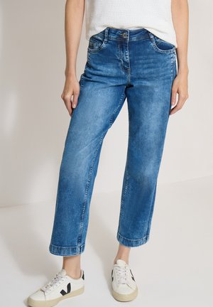 Person trägt blaue Jeans mit geradem Bein, weißes strukturiertes ärmelloses Oberteil und weiße Sneakers mit schwarzen Details, steht vor einem schlichten Hintergrund.